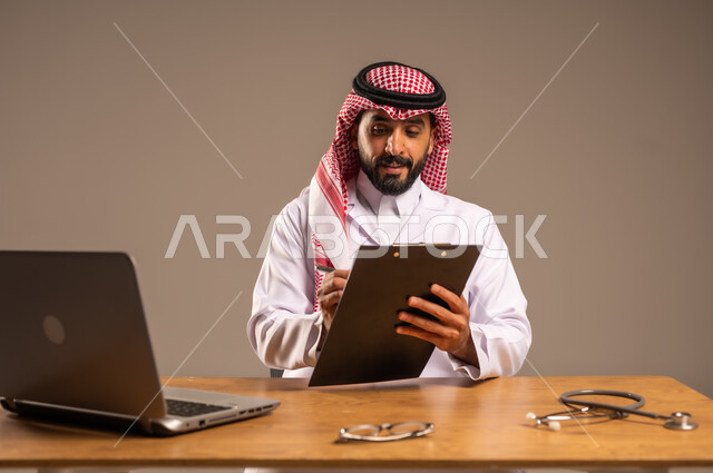 Providing services to patients, modifying and recording notes in their files, using technology in health research and development, conducting remote medical consultations, portrait of a young Saudi Arabian Gulf man wearing a medical coat and a shemagh sitting behind a desk with a laptop and an examination stethoscope in front of him, gray background