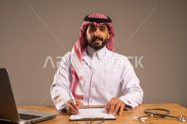 تقديم الخدمات للمرضى وتعديل وتسجيل الملاحظات في ملفاتهم، استخدام التكنولوجيا في البحث والتطوير الصحي، إجراء الاستشارات الطبية عن بعد، بورتريه لشاب عربي خليجي سعودي يرتدي المعطف الطبي والشماغ يجلس خلف المكتب وأمامه الحاسوب المحمول وسماعة الفحص، خلفية رمادية