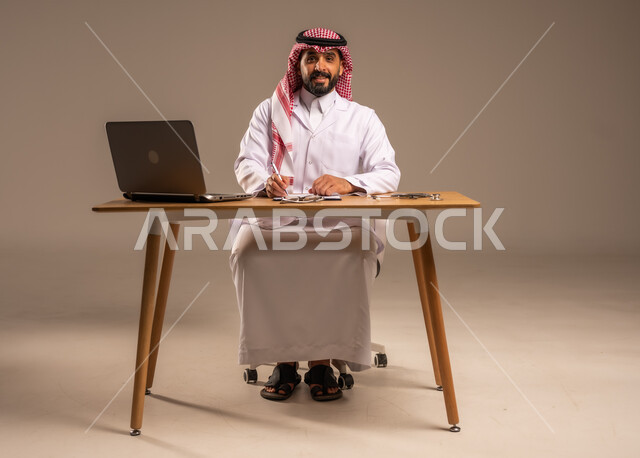 Using technology in health research and development, providing services to patients and modifying and recording notes in their files, conducting remote medical consultations, portrait of a young Saudi Arabian Gulf man wearing a medical coat and a shemagh sitting behind a desk with a laptop and an examination stethoscope in front of him, gray background