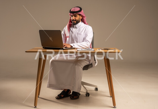 استخدام التكنولوجيا في البحث والتطوير الصحي، تقديم الخدمات للمرضى وتعديل وتسجيل الملاحظات في ملفاتهم، إجراء الاستشارات الطبية عن بعد، بورتريه لشاب عربي خليجي سعودي يرتدي المعطف الطبي والشماغ يجلس خلف المكتب وأمامه الحاسوب المحمول وسماعة الفحص، خلفية رمادية