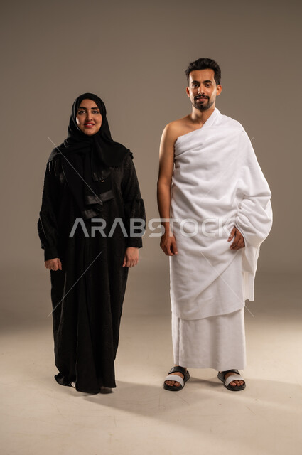 Gestures of happiness and reassurance, visiting the Holy House of Allah in Mecca, close-up portrait of a Saudi Arabian Gulf couple wearing Ihram clothes preparing to perform Hajj and Umrah rituals, getting closer to Allah through good deeds, fulfilling the fifth pillar of Islam, full-length body, beige background