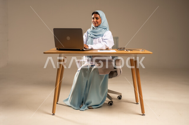 إجراء الاستشارات الطبية عن بعد، تقديم الخدمات للمرضى، استخدام التكنولوجيا في البحث والتطوير، عمل النساء في مجال الرعاية الصحية، بورتريه لامرأة عربية خليجية سعودية محجبة ترتدي المعطف الطبي تجلس خلف المكتب وأمامها الحاسوب المحمول وسماعة الفحص، خلفية رمادية