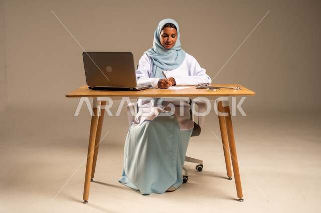 إجراء الاستشارات الطبية عن بعد، تقديم الخدمات للمرضى، استخدام التكنولوجيا في البحث والتطوير، عمل النساء في مجال الرعاية الصحية، بورتريه لامرأة عربية خليجية سعودية محجبة ترتدي المعطف الطبي تجلس خلف المكتب وأمامها الحاسوب المحمول وسماعة الفحص، خلفية رمادية