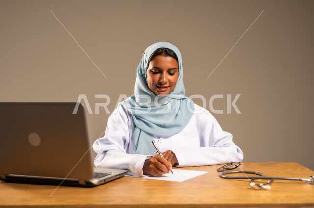 إجراء الاستشارات الطبية عن بعد، تقديم الخدمات للمرضى، استخدام التكنولوجيا في البحث والتطوير، عمل النساء في مجال الرعاية الصحية، بورتريه لامرأة عربية خليجية سعودية محجبة ترتدي المعطف الطبي تجلس خلف المكتب وأمامها الحاسوب المحمول وسماعة الفحص، خلفية رمادية