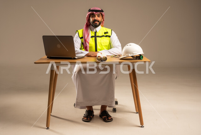 دمج العمل بالتقنيات الحديثة، بناء المملكة العربية السعودية بسواعد أبناء الوطن, بورتريه لمهندس عربي خليجي سعودي يرتدي سترة الحماية والشماغ يجلس خلف المكتب ويقوم بإدارة المشاريع، الحاسوب المحمول والمخططات الإنشائية والخوذة على الطاولة المكتبية، خلفية رمادية