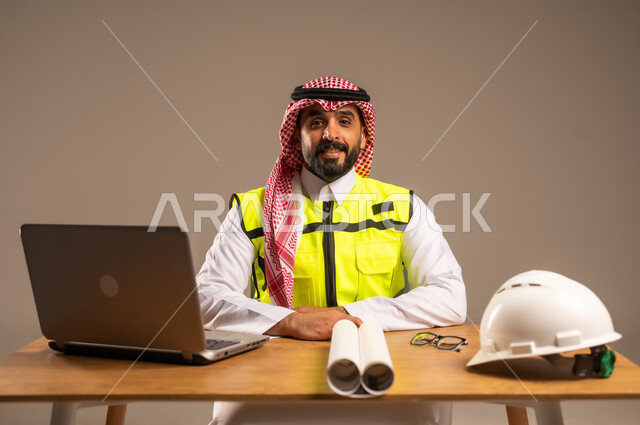 دمج العمل بالتقنيات الحديثة، بناء المملكة العربية السعودية بسواعد أبناء الوطن, بورتريه لمهندس عربي خليجي سعودي يرتدي سترة الحماية والشماغ يجلس خلف المكتب ويقوم بإدارة المشاريع، الحاسوب المحمول والمخططات الإنشائية والخوذة على الطاولة المكتبية، خلفية رمادية