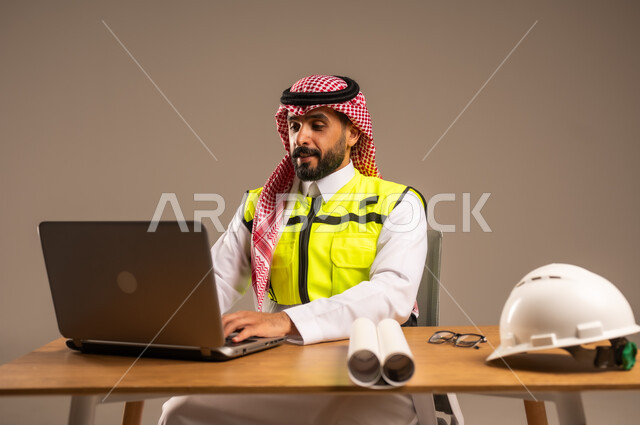دمج العمل بالتقنيات الحديثة، بناء المملكة العربية السعودية بسواعد أبناء الوطن, بورتريه لمهندس عربي خليجي سعودي يرتدي سترة الحماية والشماغ يجلس خلف المكتب ويقوم بإدارة المشاريع، الحاسوب المحمول والمخططات الإنشائية والخوذة على الطاولة المكتبية، خلفية رمادية