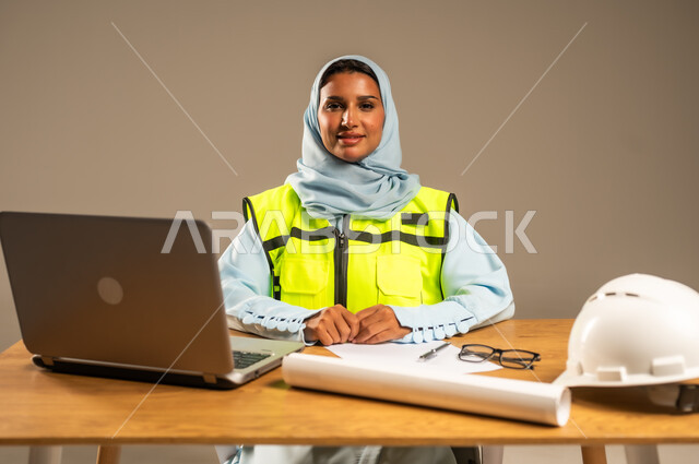 Project management using modern technologies, building the nation with the hands of its sons, portrait of a veiled Saudi Arabian Gulf female engineer wearing a protective jacket sitting behind a desk and looking at the camera with gestures of happiness, laptop, construction plans and helmet on the office table, gray background