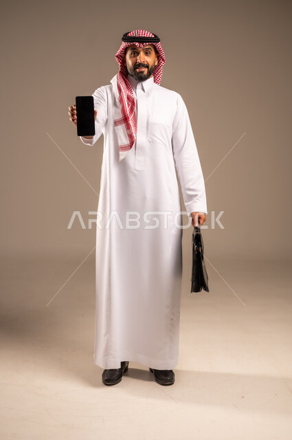 عرض شاشة سوداء فارغة، استخدام الأجهزة التقنية الحديثة في إنجاز المهام، بورتريه لرجل عربي خليجي سعودي يرتدي الثوب التقليدي والشماغ يحمل بيده الهاتف المحمول وحقيبة يد ينظر إلى الكاميرا، تحديد مواعيد اجتماعات العمل عبر الجوال، الجسم بالطول الكامل، خلفيه بيج