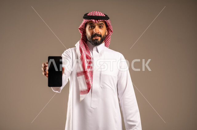 عرض شاشة سوداء فارغة، استخدام أجهزة تقنية حديثة في إنجاز المهام، بورتريه مقرب لرجل عربي خليجي سعودي يرتدي الثوب التقليدي والشماغ يحمل بيده الهاتف المحمول وحقيبة يد ينظر إلى الكاميرا، تحديد مواعيد اجتماعات العمل عبر الجوال، دمج التكنولوجيا مع الأعمال، خلفيه بيج