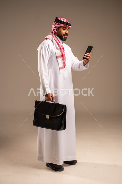 استخدام الأجهزة التقنية الحديثة في إنجاز المهام، بورتريه لرجل عربي خليجي سعودي يرتدي الثوب التقليدي والشماغ يحمل بيده الهاتف المحمول وحقيبة يد، تحديد مواعيد اجتماعات العمل عبر الجوال، دمج التكنولوجيا مع الأعمال، الجسم بالطول الكامل، خلفيه بيج