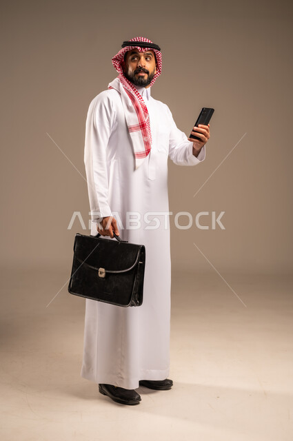 دمج التكنولوجيا مع الأعمال، استخدام الأجهزة التقنية الحديثة في إنجاز المهام، بورتريه لرجل عربي خليجي سعودي يرتدي الثوب التقليدي والشماغ يحمل بيده الهاتف المحمول وحقيبة يد ينظر إلى شيء ما، تحديد مواعيد اجتماعات العمل عبر الجوال، الجسم بالطول الكامل، خلفيه بيج