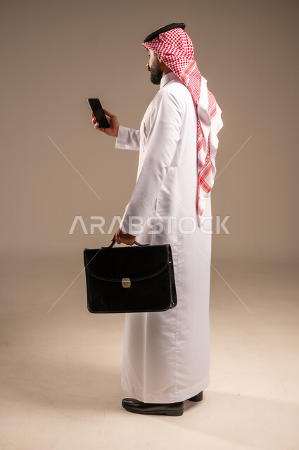 دمج التكنولوجيا مع الأعمال، استخدام الأجهزة التقنية الحديثة في إنجاز المهام، بورتريه من الجانب لرجل عربي خليجي سعودي يرتدي الثوب التقليدي والشماغ يحمل بيده الهاتف المحمول وحقيبة يد، تحديد مواعيد اجتماعات العمل عبر الجوال، الجسم بالطول الكامل، خلفيه بيج