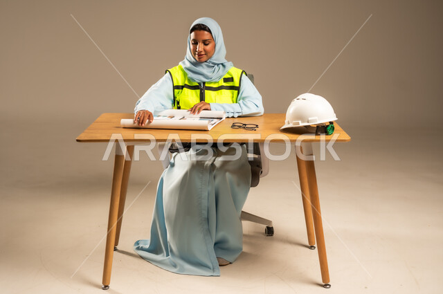 مفهوم الهندسة والإنشاءات المعمارية، بورتريه لمهندسة عربية خليجية سعودية محجبة ترتدي السترة الخاصة بالعمل تجلس خلف المكتب وأمامها مخططات هندسية وخوذة الحماية، تعديل وتدقيق مخطوطات البناء، معاينه تطبيق الخطط والإشراف على سير المشاريع، خلفية رمادية