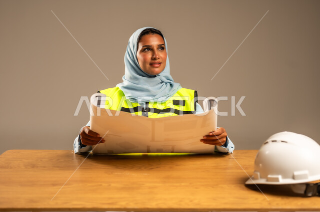 مفهوم الهندسة والإنشاءات المعمارية، بورتريه لمهندسة عربية خليجية سعودية محجبة ترتدي السترة الخاصة بالعمل تجلس خلف المكتب وأمامها مخططات هندسية وخوذة الحماية، تعديل وتدقيق مخطوطات البناء، معاينه تطبيق الخطط والإشراف على سير المشاريع، خلفية رمادية