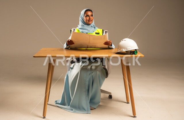 مفهوم الهندسة والإنشاءات المعمارية، بورتريه لمهندسة عربية خليجية سعودية محجبة ترتدي السترة الخاصة بالعمل تجلس خلف المكتب وأمامها مخططات هندسية وخوذة الحماية، تعديل وتدقيق مخطوطات البناء، معاينه تطبيق الخطط والإشراف على سير المشاريع، خلفية رمادية