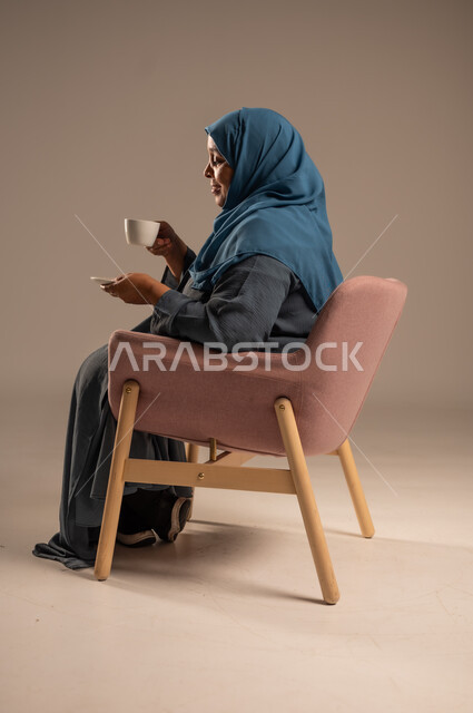  أصالة عادات وتقاليد المجتمع السعودي, كرم الضيافة وحسن الاستقبال في المملكة, بورتريه لامراة كبيرة بالسن عربية خليجية سعودية ترتدي العباءة والحجاب تجلس على كرسي مريح تحمل بيدها كوب من القهوة، الاهتمام بالمظهر الخارجي، خلفية ملونة