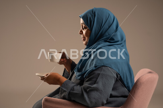 Authenticity of Saudi society customs and traditions, hospitality and good reception in the Kingdom, portrait of an elderly Saudi Arabian Gulf woman wearing an abaya and hijab sitting on a comfortable chair holding a cup of coffee in her hand, interest in outward appearance, colorful background