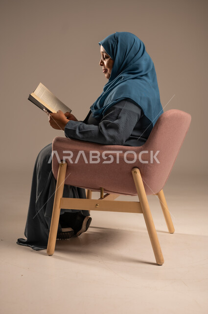 Enjoying reading books, stories and novels, self-learning culture, practicing the hobby of reading and studying, the concept of literacy, spending free time learning and culture, portrait of a veiled Saudi Arabian Gulf woman wearing an abaya sitting on a comfortable chair holding a book in her hand, beige background