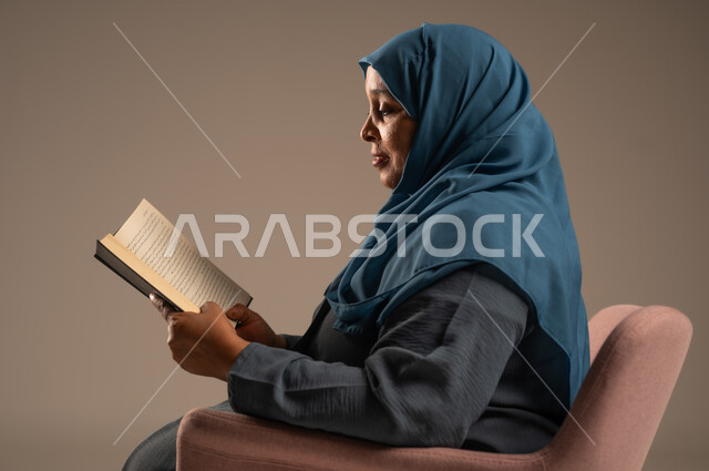 الاستمتاع بقراءة الكتب والقصص والروايات, ثقافة التعلم الذاتي، ممارسة هواية القراءة والمطالعة, مفهوم محو الأمية، قضاء وقت الفراغ بالتعلم والثقافة، بورتريه لامراة عربية خليجية سعودية محجبة ترتدي العباءة تجلس على كرسي مريح تحمل بيدها كتاب، خلفية بيج