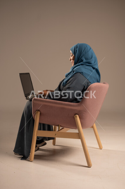 ادارة الأعمال عن بعد، استخدام الأجهزة الحديثة والمتطورة، بورتريه لامرأة كبيرة بالسن عربية خليجية سعودية ترتدي العباءة والحجاب تقوم باستخدام الحاسوب المحمول، دمج التكنولوجيا بالعمل، إنجاز المهام المطلوبة عبر اللابتوب، مهن ووظائف نسائية، خلفية ملونة