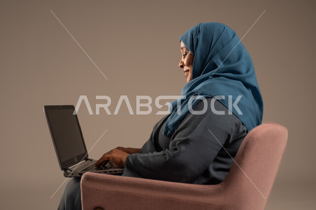 ادارة الأعمال عن بعد، استخدام الأجهزة الحديثة والمتطورة، بورتريه لامرأة كبيرة بالسن عربية خليجية سعودية ترتدي العباءة والحجاب تقوم باستخدام الحاسوب المحمول، دمج التكنولوجيا بالعمل، إنجاز المهام المطلوبة عبر اللابتوب، مهن ووظائف نسائية، خلفية ملونة