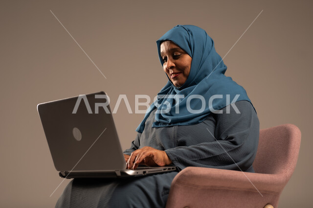 ادارة الأعمال عن بعد، استخدام الأجهزة الحديثة والمتطورة، بورتريه لامرأة كبيرة بالسن عربية خليجية سعودية ترتدي العباءة والحجاب تقوم باستخدام الحاسوب المحمول، دمج التكنولوجيا بالعمل، إنجاز المهام المطلوبة عبر اللابتوب، مهن ووظائف نسائية، خلفية ملونة