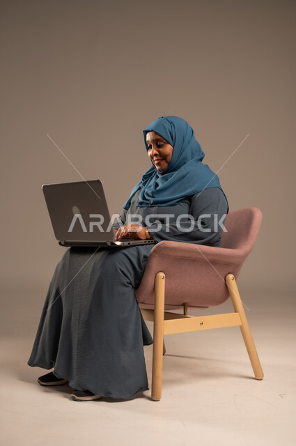ادارة الأعمال عن بعد، استخدام الأجهزة الحديثة والمتطورة، بورتريه لامرأة كبيرة بالسن عربية خليجية سعودية ترتدي العباءة والحجاب تقوم باستخدام الحاسوب المحمول، دمج التكنولوجيا بالعمل، إنجاز المهام المطلوبة عبر اللابتوب، مهن ووظائف نسائية، خلفية ملونة