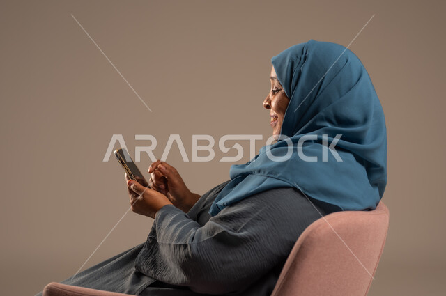 تصفح التطبيقات وبرامج التواصل الاجتماعي، تبادل الأحاديث والأخبار مع الأهل والأصدقاء، بورتريه لإمرأة عربية خليجية سعودية ترتدي العباءة والحجاب تجلس على كرسي مريح تمسك بيدها الهاتف المحمول، استخدام الأجهزة والتقنيات الحديثة والمتطورة، خلفية ملونة