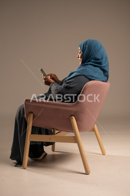 تصفح التطبيقات وبرامج التواصل الاجتماعي، تبادل الأحاديث والأخبار مع الأهل والأصدقاء، بورتريه لإمرأة عربية خليجية سعودية ترتدي العباءة والحجاب تجلس على كرسي مريح تمسك بيدها الهاتف المحمول، استخدام الأجهزة والتقنيات الحديثة والمتطورة، خلفية ملونة