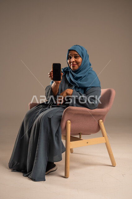 تصفح التطبيقات وبرامج التواصل الاجتماعي، تبادل الأحاديث والأخبار مع الأهل والأصدقاء، بورتريه لإمرأة عربية خليجية سعودية ترتدي العباءة والحجاب تجلس على كرسي مريح تمسك بيدها الهاتف المحمول، استخدام الأجهزة والتقنيات الحديثة والمتطورة، خلفية ملونة