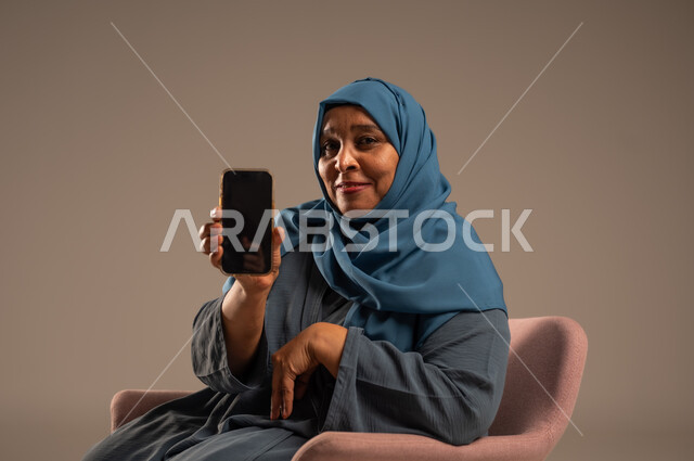 تصفح التطبيقات وبرامج التواصل الاجتماعي، تبادل الأحاديث والأخبار مع الأهل والأصدقاء، بورتريه لإمرأة عربية خليجية سعودية ترتدي العباءة والحجاب تجلس على كرسي مريح تمسك بيدها الهاتف المحمول، استخدام الأجهزة والتقنيات الحديثة والمتطورة، خلفية ملونة