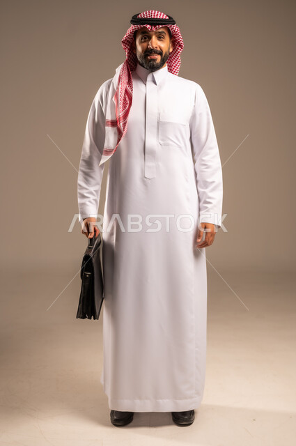 مفهوم إداة الأعمال والمشاريع، الاستعداد للذهاب إلى العمل، بورتريه لرجل عربي خليجي سعودي يرتدي الثوب التقليدي والشماغ يحمل بيده حقيبة يد يقف باستقامة ينظر إلى الكاميرا بإيماءات الثقة بالنفس، الأناقة والاهتمام بالمظهر الخارجي، الجسم بالطول الكامل، خلفيه بيج
