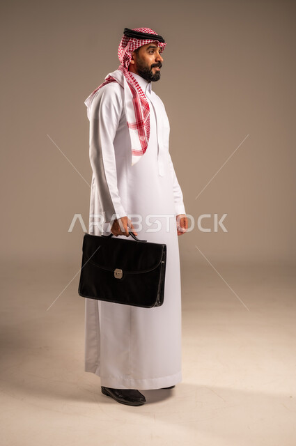 الذهاب إلى العمل بنشاط، بورتريه من الجانب لرجل عربي خليجي سعودي يرتدي الثوب التقليدي والشماغ يحمل بيده حقيبة يد يمشي إلى مكان ما ينظر أمامه بإيماءات الثقة بالنفس، الاهتمام بالمظهر الخارجي، مفهوم إداة الأعمال والمشاريع، الجسم بالطول الكامل، خلفيه بيج