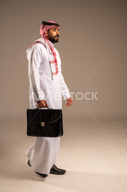 الذهاب إلى العمل بنشاط، بورتريه من الجانب لرجل عربي خليجي سعودي يرتدي الثوب التقليدي والشماغ يحمل بيده حقيبة يد يمشي إلى مكان ما ينظر أمامه بإيماءات الثقة بالنفس، الاهتمام بالمظهر الخارجي، مفهوم إداة الأعمال والمشاريع، الجسم بالطول الكامل، خلفيه بيج