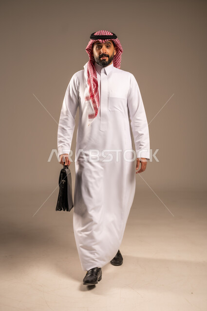الأناقة والاهتمام بالمظهر الخارجي، الذهاب إلى العمل بنشاط، بورتريه لرجل عربي خليجي سعودي يرتدي الثوب التقليدي والشماغ يحمل بيده حقيبة يد يمشي إلى مكان ما ينظر إلى الكاميرا بإيماءات الثقة بالنفس، مفهوم إداة الأعمال والمشاريع، الجسم بالطول الكامل، خلفيه بيج