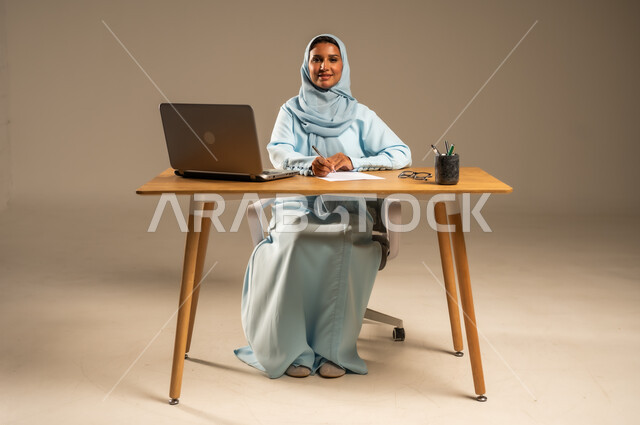 إنجاز المهام المطلوبة عبر جهاز الحاسوب المحمول، وظائف ومهن مكتبية إدارية نسائية في السعودية، دمج العمل مع التكنولوجيا الحديثة، بورتريه لامرأة عربية خليجية سعودية محجبة تجلس خلف المكتب وتقوم بمتابعة عملها أونلاين، مفهوم العمل عن بعد عبر الانترنت، خلفية رمادية