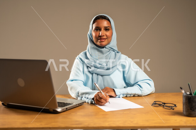 إنجاز المهام المطلوبة عبر جهاز الحاسوب المحمول، وظائف ومهن مكتبية إدارية نسائية في السعودية، دمج العمل مع التكنولوجيا الحديثة، بورتريه لامرأة عربية خليجية سعودية محجبة تجلس خلف المكتب وتقوم بمتابعة عملها أونلاين، مفهوم العمل عن بعد عبر الانترنت، خلفية رمادية