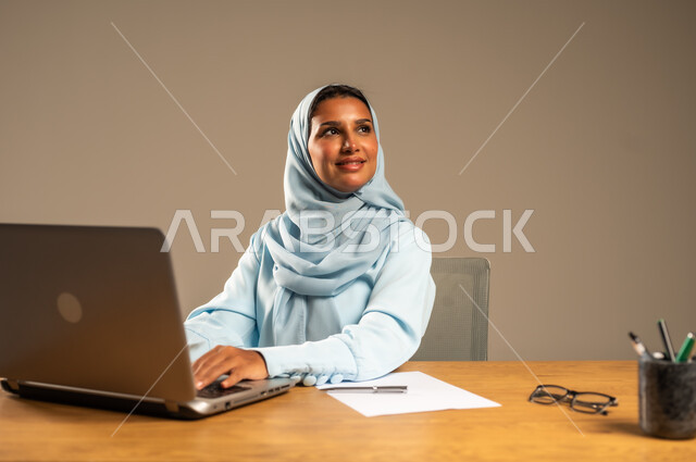 إنجاز المهام المطلوبة عبر جهاز الحاسوب المحمول، وظائف ومهن مكتبية إدارية نسائية في السعودية، دمج العمل مع التكنولوجيا الحديثة، بورتريه لامرأة عربية خليجية سعودية محجبة تجلس خلف المكتب وتقوم بمتابعة عملها أونلاين، مفهوم العمل عن بعد عبر الانترنت، خلفية رمادية