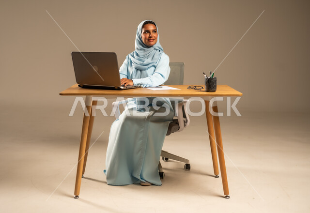 إنجاز المهام المطلوبة عبر جهاز الحاسوب المحمول، وظائف ومهن مكتبية إدارية نسائية في السعودية، دمج العمل مع التكنولوجيا الحديثة، بورتريه لامرأة عربية خليجية سعودية محجبة تجلس خلف المكتب وتقوم بمتابعة عملها أونلاين، مفهوم العمل عن بعد عبر الانترنت، خلفية رمادية