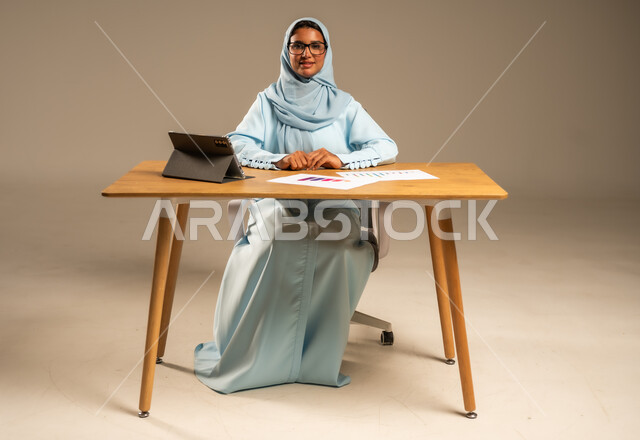 عمل النساء في المجالات الإدارية المكتبية في السعودية، مفهوم العمل عن بعد عبر الانترنت، إنجاز المهام المطلوبة أونلاين، دمج الأعمال مع التكنولوجيا والتقنيات الحديثة، بورتريه لامرأة عربية خليجية سعودية محجبة تجلس خلف المكتب وتعمل عبر الجهاز اللوحي، خلفية رمادية