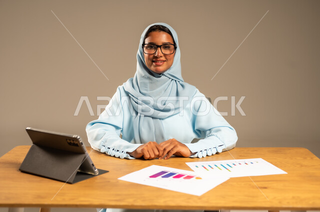 عمل النساء في المجالات الإدارية المكتبية في السعودية، مفهوم العمل عن بعد عبر الانترنت، إنجاز المهام المطلوبة أونلاين، دمج الأعمال مع التكنولوجيا والتقنيات الحديثة، بورتريه لامرأة عربية خليجية سعودية محجبة تجلس خلف المكتب وتعمل عبر الجهاز اللوحي، خلفية رمادية