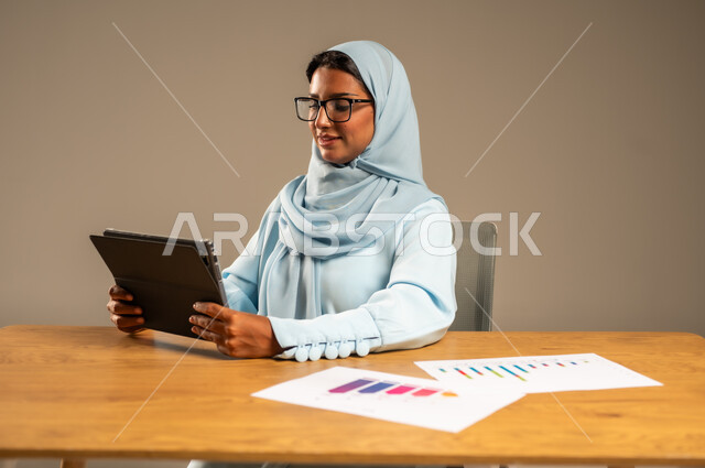 عمل النساء في المجالات الإدارية المكتبية في السعودية، مفهوم العمل عن بعد عبر الانترنت، إنجاز المهام المطلوبة أونلاين، دمج الأعمال مع التكنولوجيا والتقنيات الحديثة، بورتريه لامرأة عربية خليجية سعودية محجبة تجلس خلف المكتب وتعمل عبر الجهاز اللوحي، خلفية رمادية