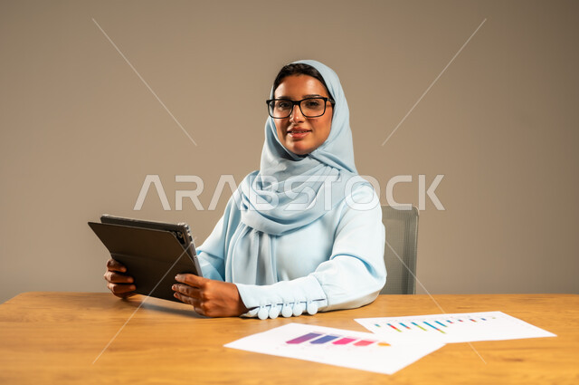 مفهوم العمل عن بعد عبر الانترنت، مهن ووظائف مكتبية إدارية نسائية في السعودية، إنجاز المهام المطلوبة أونلاين، دمج العمل مع التكنولوجيا الحديثة، بورتريه لامرأة عربية خليجية سعودية محجبة تجلس خلف المكتب وتقوم بمتابعة عملها عبر الجهاز اللوحي، خلفية رمادية
