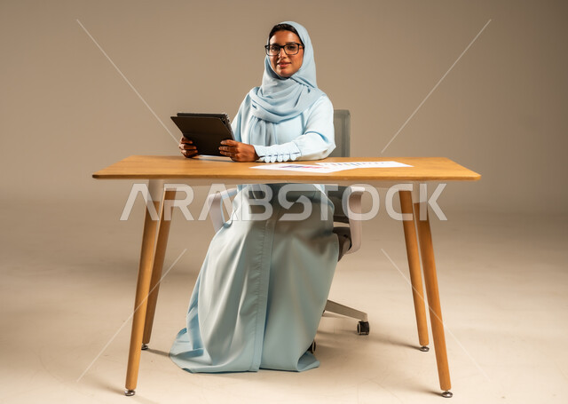 عمل النساء في المجالات الإدارية المكتبية في السعودية، مفهوم العمل عن بعد عبر الانترنت، إنجاز المهام المطلوبة أونلاين، دمج الأعمال مع التكنولوجيا والتقنيات الحديثة، بورتريه لامرأة عربية خليجية سعودية محجبة تجلس خلف المكتب وتعمل عبر الجهاز اللوحي، خلفية رمادية