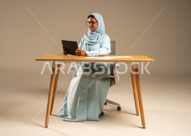مفهوم العمل عن بعد عبر الانترنت، مهن ووظائف مكتبية إدارية نسائية في السعودية، إنجاز المهام المطلوبة أونلاين، دمج العمل مع التكنولوجيا الحديثة، بورتريه لامرأة عربية خليجية سعودية محجبة تجلس خلف المكتب وتقوم بمتابعة عملها عبر الجهاز اللوحي، خلفية رمادية