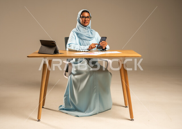 دمج الأعمال مع التكنولوجيا الحديثة، استخدام الهاتف المحمول في العمل أونلاين، مهن ووظائف مكتبية للنساء في السعودية، مفهوم العمل عن بعد عبر الانترنت، بورتريه لامرأة عربية خليجية سعودية محجبة تجلس خلف المكتب وأمامها الجهاز اللوحي وعدد من الأوراق، خلفية رمادية