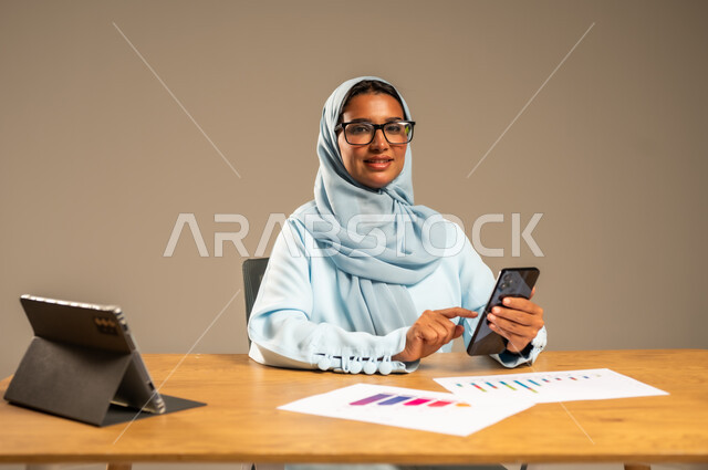 دمج الأعمال مع التكنولوجيا الحديثة، استخدام الهاتف المحمول في العمل أونلاين، مهن ووظائف مكتبية للنساء في السعودية، مفهوم العمل عن بعد عبر الانترنت، بورتريه لامرأة عربية خليجية سعودية محجبة تجلس خلف المكتب وأمامها الجهاز اللوحي وعدد من الأوراق، خلفية رمادية