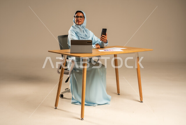 استخدام الهاتف المحمول في العمل أونلاين، مهن ووظائف مكتبية للنساء في السعودية، مفهوم العمل عن بعد عبر الانترنت، دمج الأعمال مع التكنولوجيا الحديثة، بورتريه لامرأة عربية خليجية سعودية محجبة تجلس خلف المكتب وأمامها الجهاز اللوحي وعدد من الأوراق، خلفية رمادية