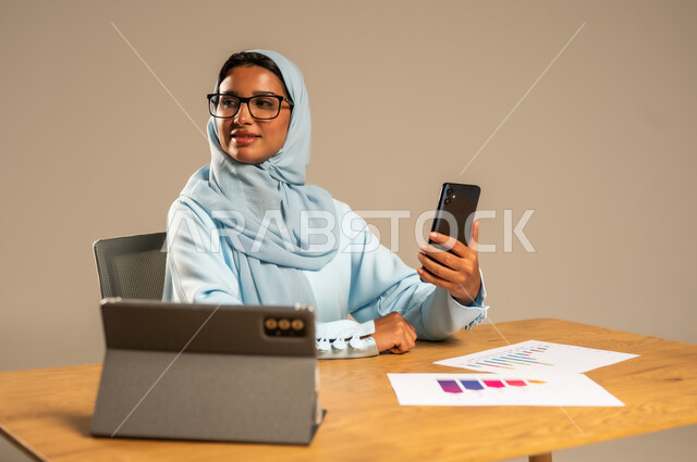 استخدام الهاتف المحمول في العمل أونلاين، مهن ووظائف مكتبية للنساء في السعودية، مفهوم العمل عن بعد عبر الانترنت، دمج الأعمال مع التكنولوجيا الحديثة، بورتريه لامرأة عربية خليجية سعودية محجبة تجلس خلف المكتب وأمامها الجهاز اللوحي وعدد من الأوراق، خلفية رمادية